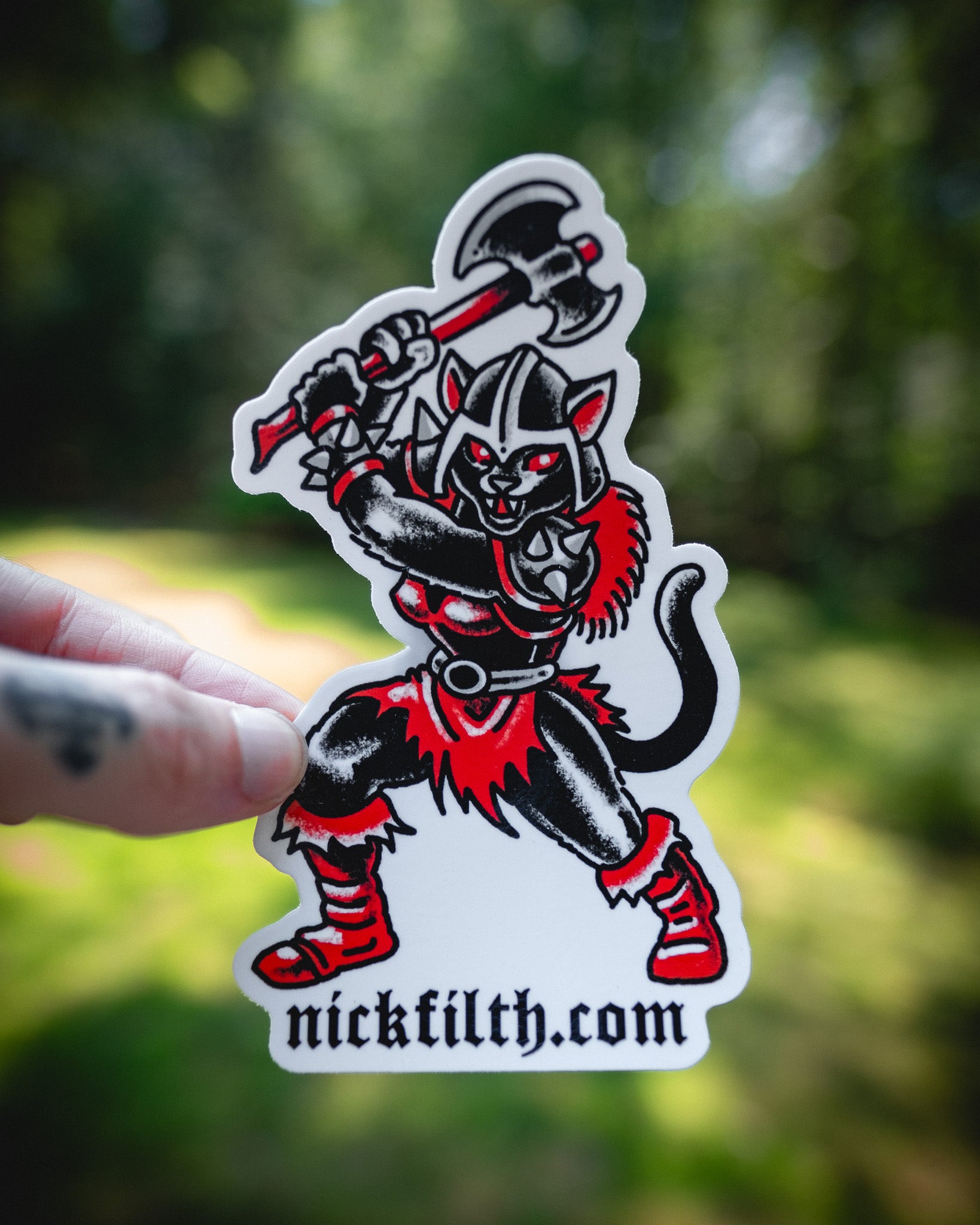Sticker of a black and red character with 'nickfilth.com' text held against a blurred green background
