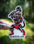 Sticker of a black and red character with 'nickfilth.com' text held against a blurred green background