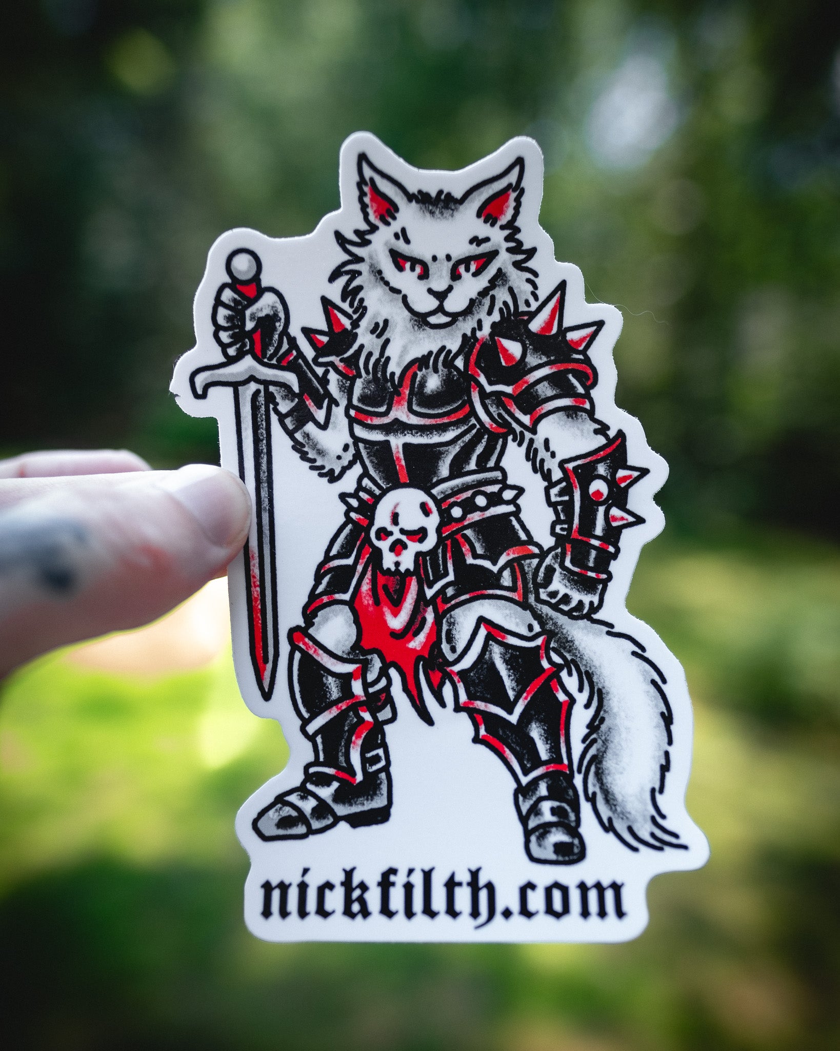 Sticker of a warrior cat with armor and a sword, held against a blurred green background.