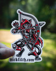 Sticker of a hooded figure with a bow and arrow held by a hand, against a blurred natural background.