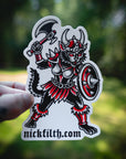 Sticker of a warrior creature with a shield and axe, held against a blurred natural background.