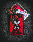 Enamel pin with 'Filth' design on a dark textured background